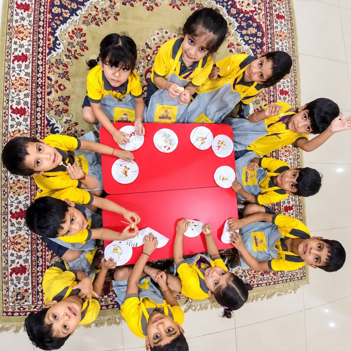 Kidzee Ramnagar children in aprons around a red table, looking up from their fruit activity
